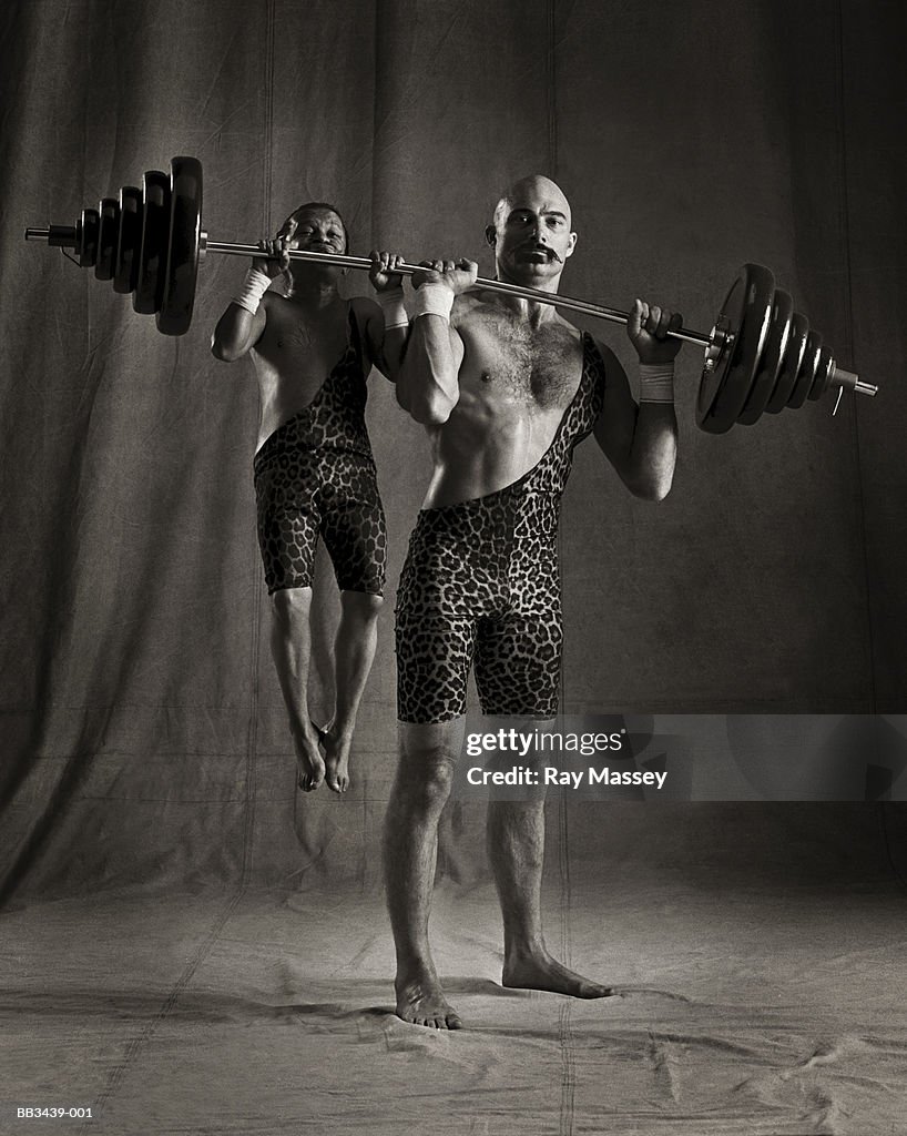 Circus dwarf hanging from barbell lifted by strongman (tinted B&W)