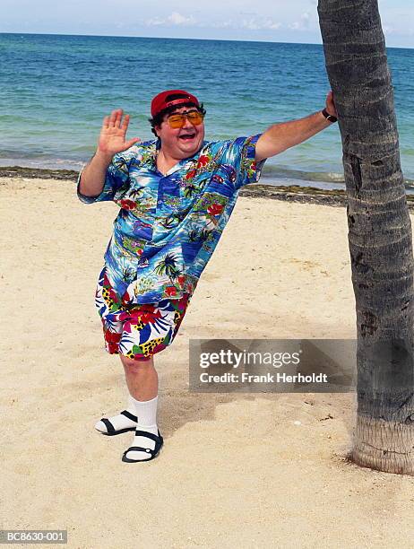 overweight man standing on beach, leaning on palm tree, waving - fat man on beach stock pictures, royalty-free photos & images