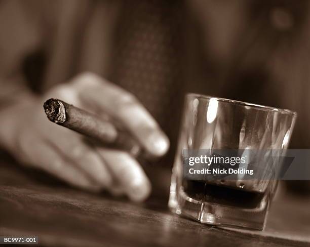 man's hand holding cigar, resting beside glass of alcohol (toned b&w) - bar drink establishment stock pictures, royalty-free photos & images