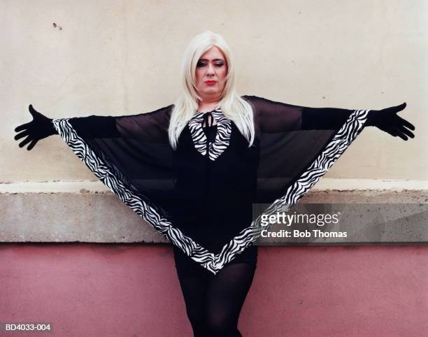 Man in drag posing by wall, arms oustretched, winking
