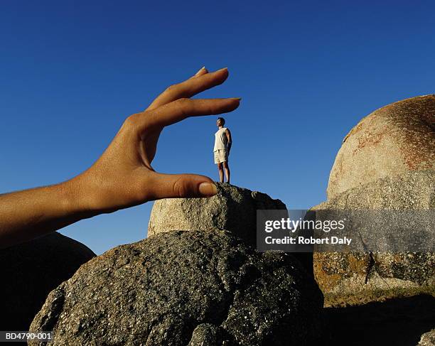 man standing on rock, hand sizing him in foreground (composite) - paradox stock pictures, royalty-free photos & images