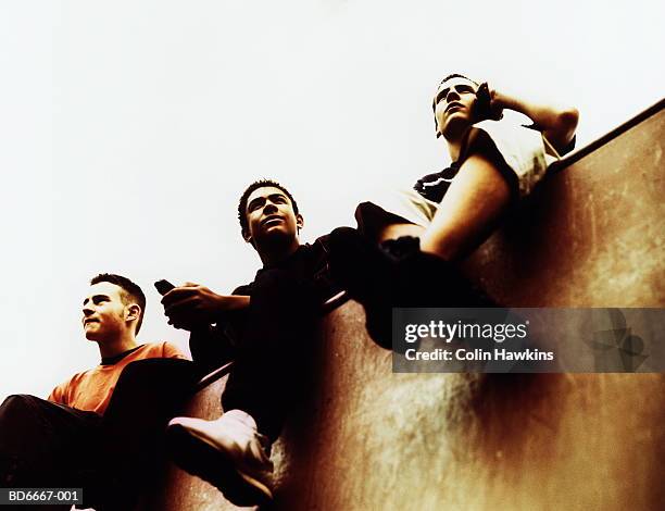 three teenage boys (15-17) sitting on wall, using mobile phones - 3 teenagers mobile outdoors stock-fotos und bilder