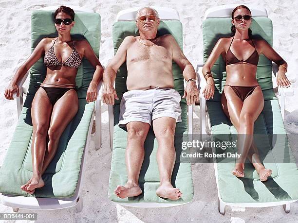 mature man and two young women on sun loungers on beach - gebruind stockfoto's en -beelden