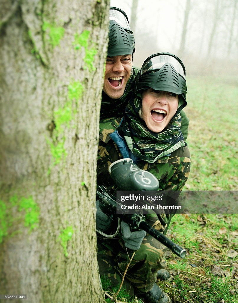 Couple paintballing, hiding behind tree