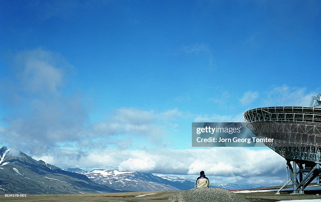 Man sitting near satellite dish, rear view