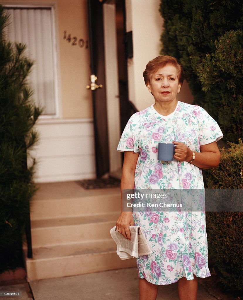 Woman standing in front of house, holding cup and newspaper, portrait