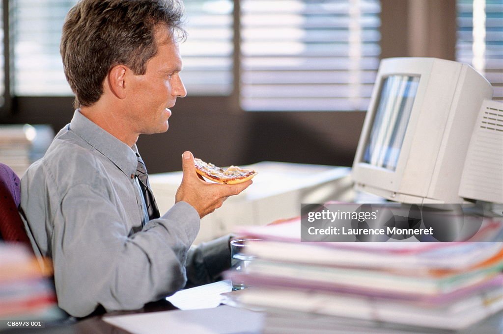 Male excutive sitting at computer in office, eating slice of pizza