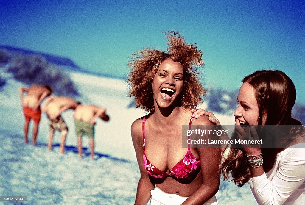 Two women on beach, laughing at men showing buttocks