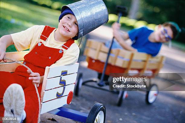 boy with bucket on head riding in cart; second boy in wagon behind him - fahrspaß stock-fotos und bilder