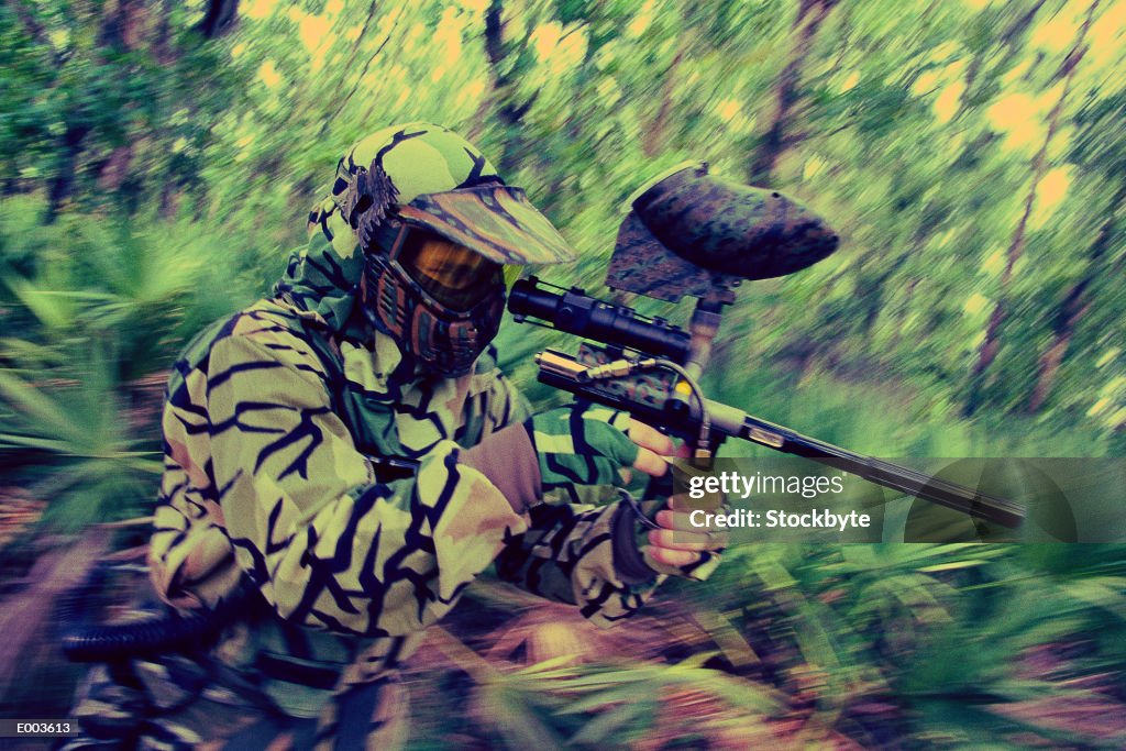 Man in fatigues playing paintball, aiming at target in woods