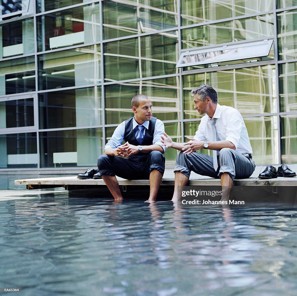 Two businessmen sitting outdoors, feet in pool of water