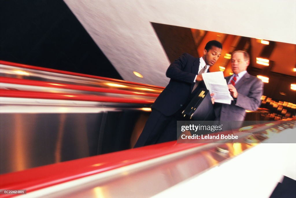 Businessmen descending escalator, reviewing papers