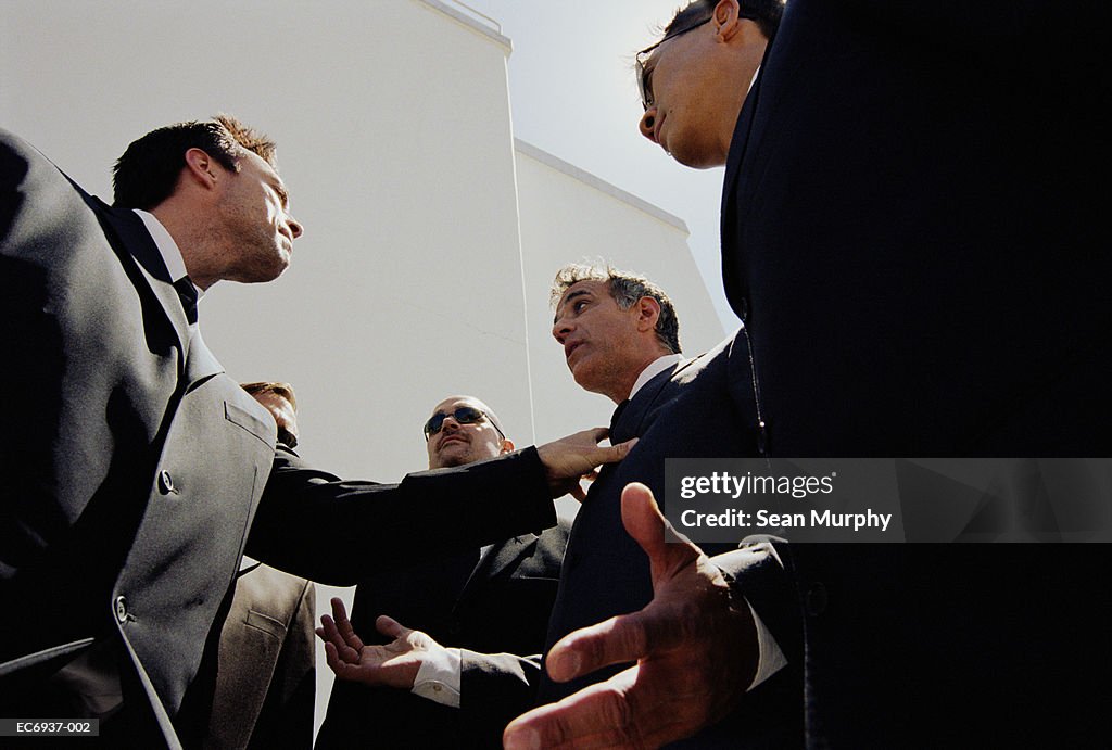 Two men in suits arguing, surrounded by bodyguards