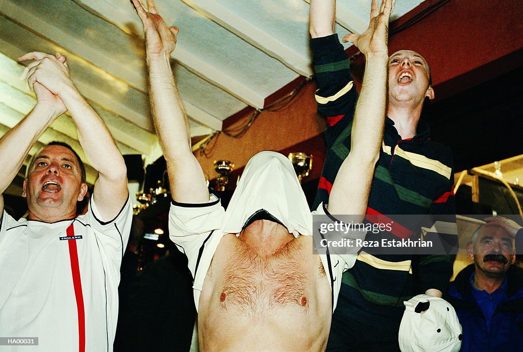 Cheering football fan covering face with shirt at pub, arms raised