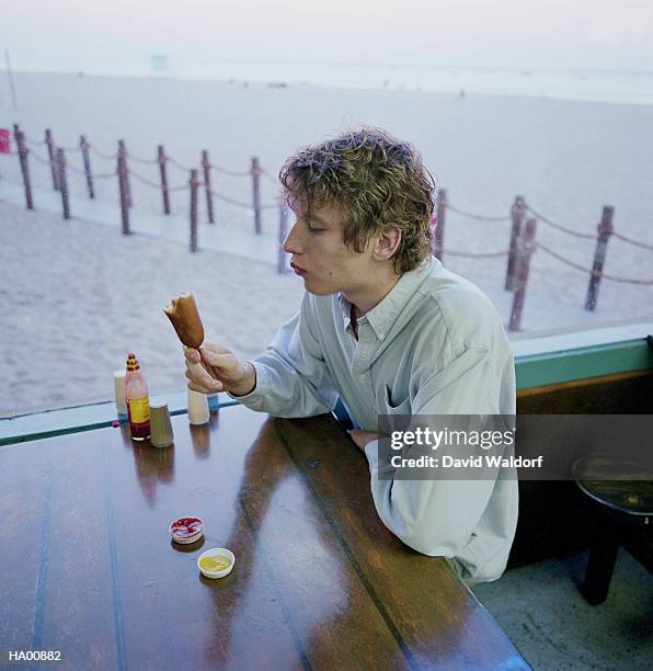 young man eating corndog, ketchup and mustard containers on table - waldorf fotografías e imágenes de stock