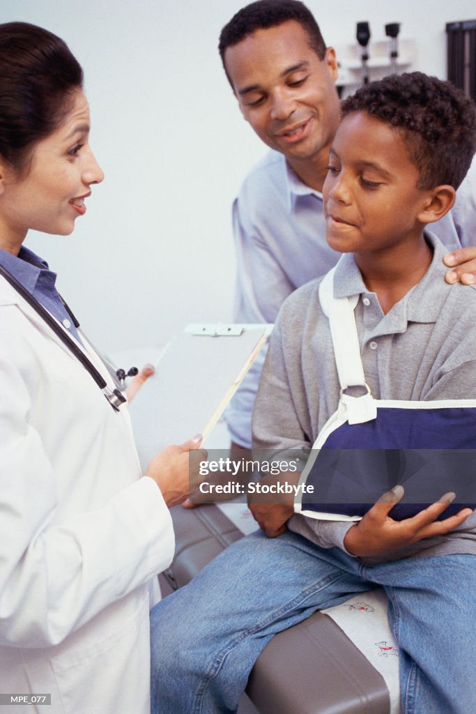 Doctor looking at boy with arm in sling; father in background