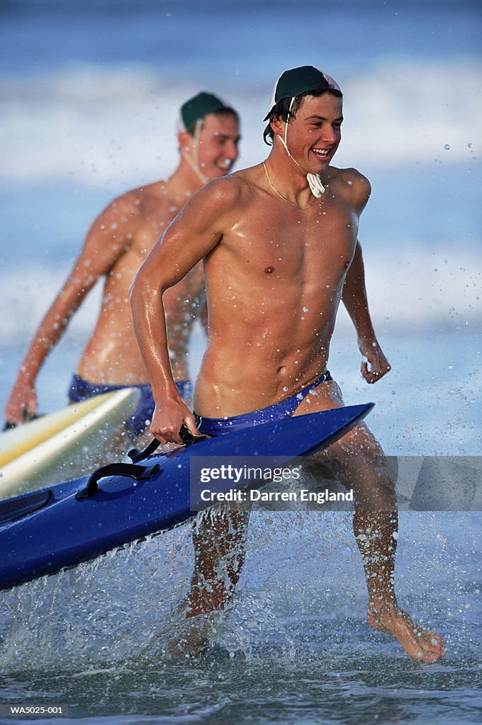 Two male lifeguards running in surf, carrying paddle boards