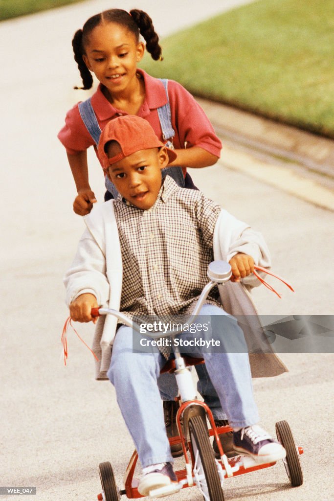 Boy riding tricycle; girl running behind him