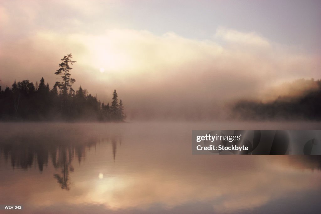 Mist rising above lake, tree-lined shore in distance