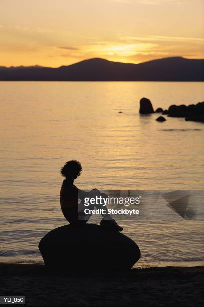 silhouette of woman sitting on rock at lakeshore at sunset - at fotografías e imágenes de stock