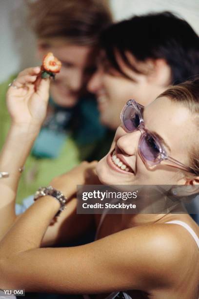 woman holding strawberry with bite out of it; couple in background - in between stock-fotos und bilder