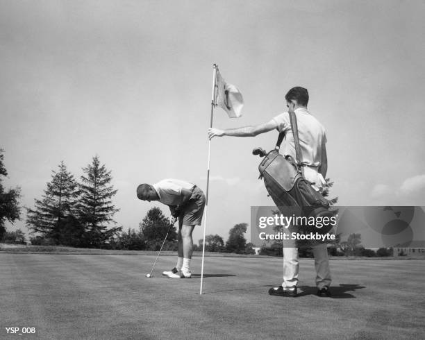 man making golf shot while caddie stands holding flag - golf caddy stockfoto's en -beelden