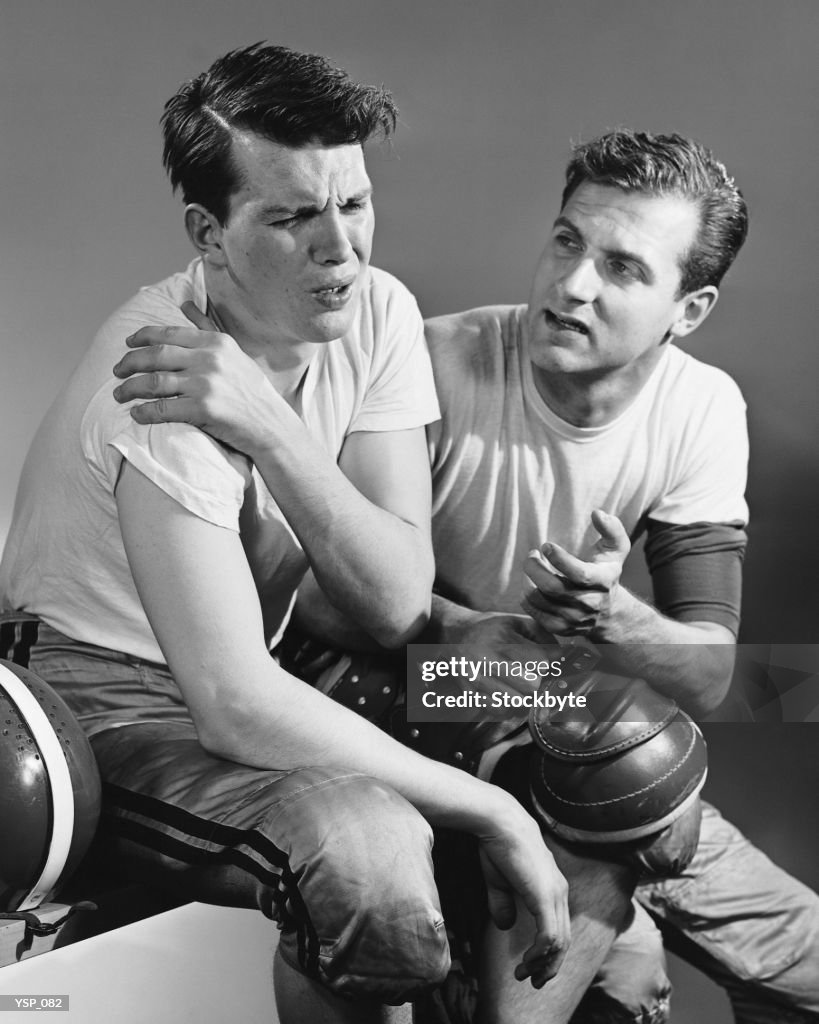 Football player holding sore shoulder; trainer sitting beside him
