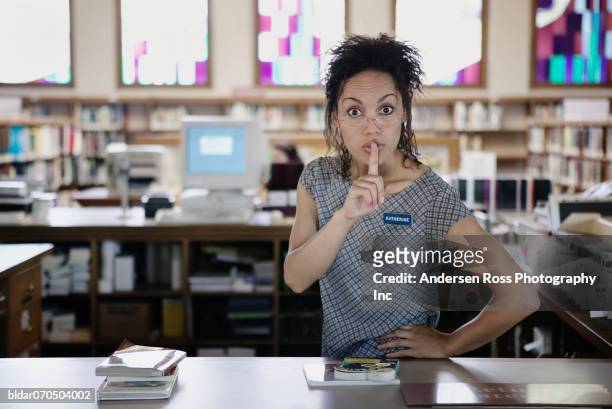portrait of a young female librarian standing in a library with a finger on her lips - stilte stockfoto's en -beelden