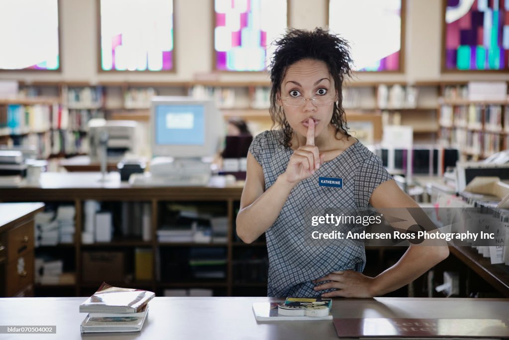 Portrait of a young female librarian standing in a library with a finger on her lips