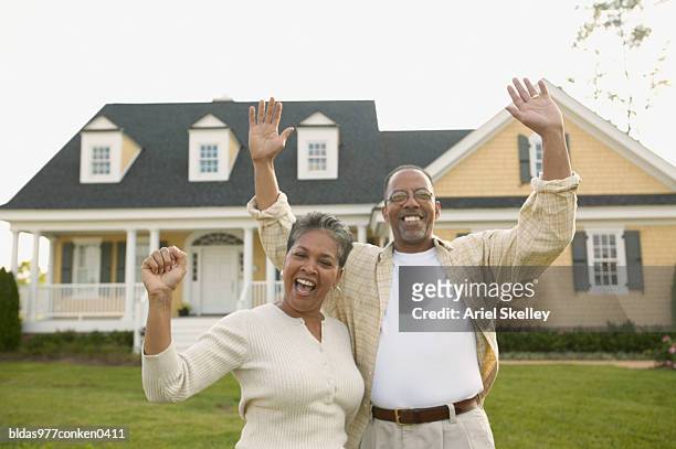portrait of a mature couple standing together with their arms raised in front of a house - ariel skelley stock-fotos und bilder