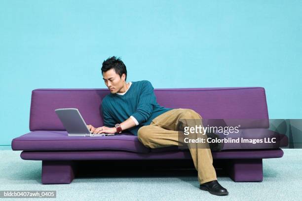 young man lying on a couch operating a laptop - asian man sitting casual imagens e fotografias de stock