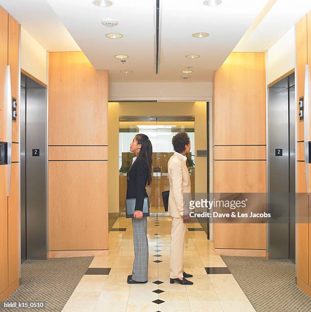 side profile of two businesswomen waiting outside an elevator - dave and les jacobs stock-fotos und bilder