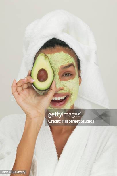 portrait of a young woman wearing a facial mask holding a slice of avocado - clay mask stock pictures, royalty-free photos & images