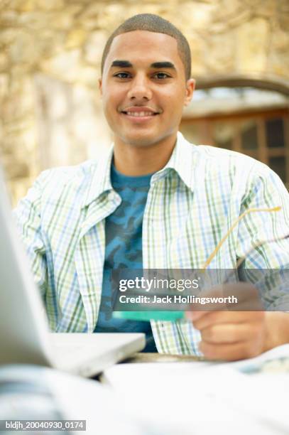 young man sitting at a table in front of a laptop holding a credit card - card table stock pictures, royalty-free photos & images