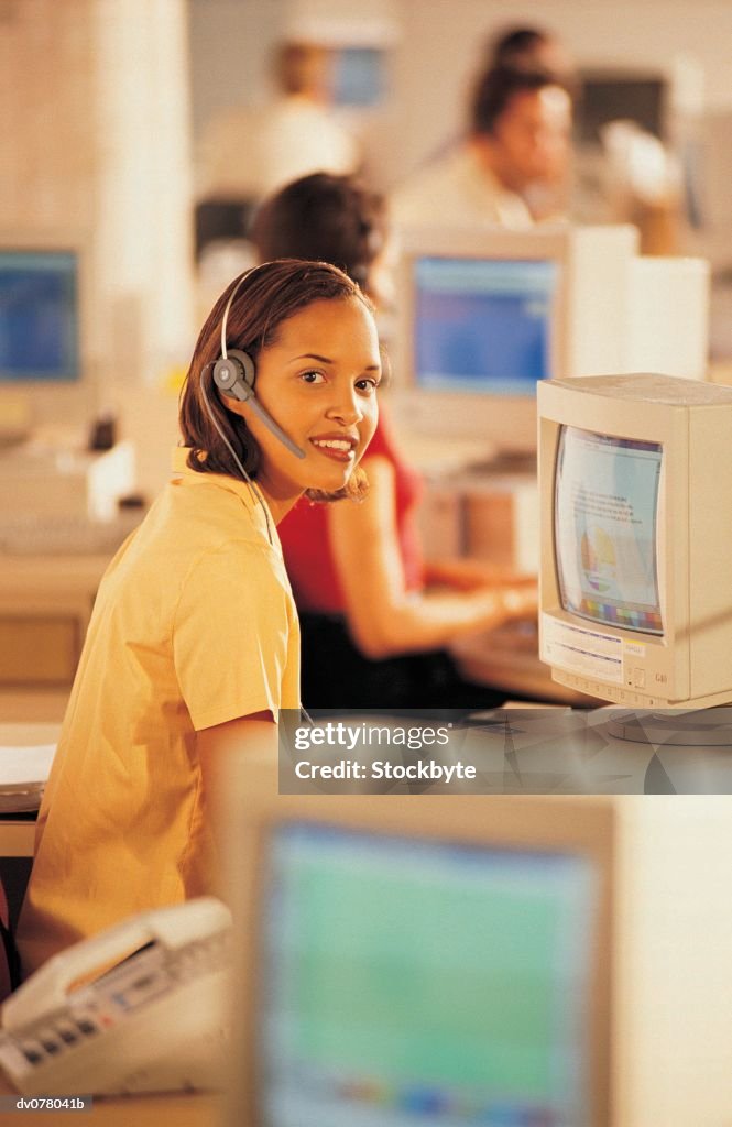 Woman seated at computer workstation, wearing telephone headset, smiling