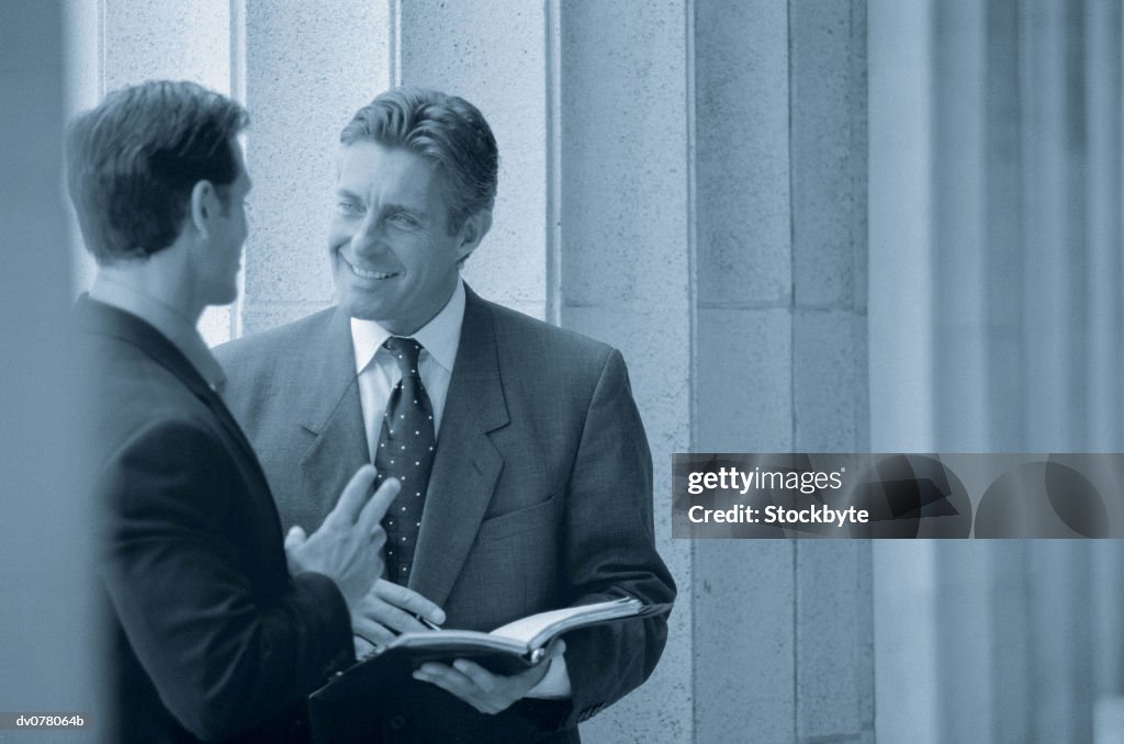 Two men standing by columns, talking; one man holding open book