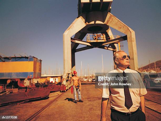 mature businessman standing in a harbour wearing sunglasses and looking sideways with a docker standing in the background - docker stock pictures, royalty-free photos & images