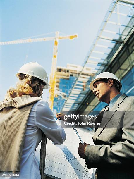 businessman and businesswoman wearing hard hats reading a blueprint on a building site - low angle view of two businessmen standing face to face outdoors stock pictures, royalty-free photos & images