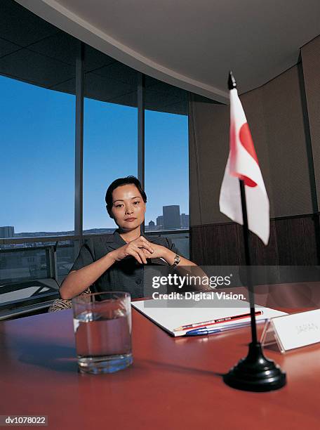 portrait of a young businesswoman sitting at a conference table by a japanese flag with her hands together - upper midtown manhattan stock pictures, royalty-free photos & images