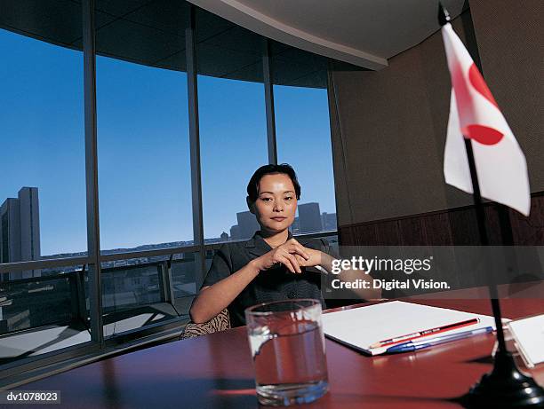 portrait of a young businesswoman sitting at a conference table by a japanese flag with her hands together - upper midtown manhattan stock pictures, royalty-free photos & images