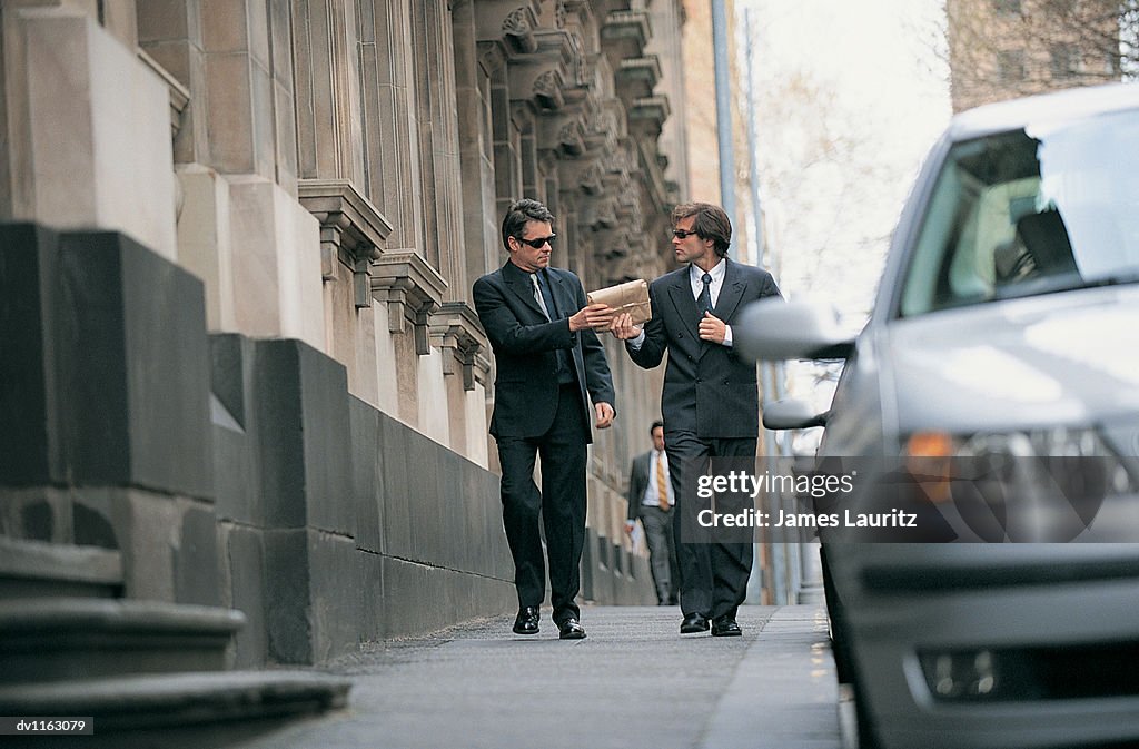 Suspicious Businessman Walking on a City Pavement Passing a Parcel to a Businessman Walking Next to Him
