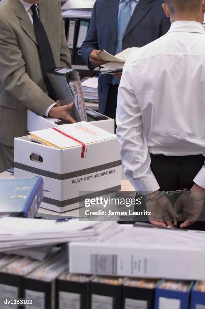 businessman standing in an office wearing handcuffs and businessmen searching through folders and documents - police scrutiny stock pictures, royalty-free photos & images