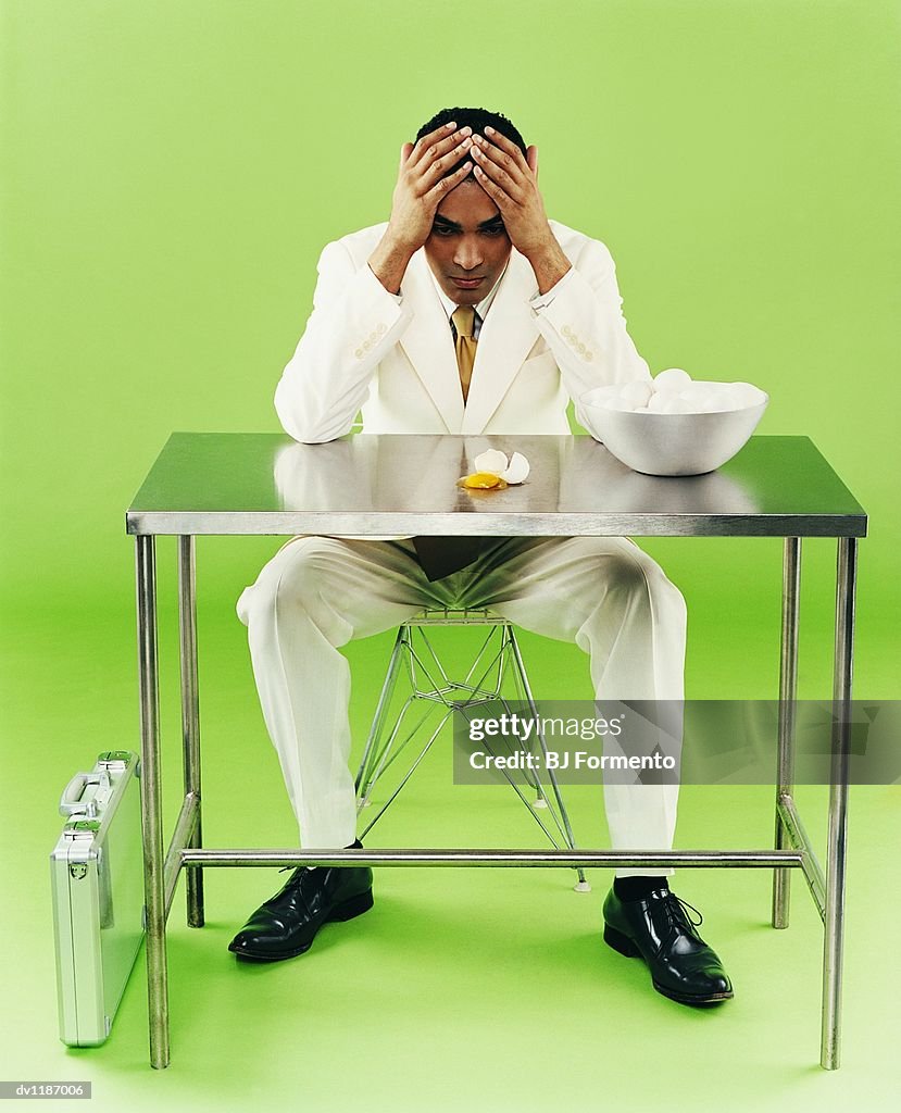 Businessman Looking Down at a Broken Egg Sitting on Chair Behind a Metallic Desk