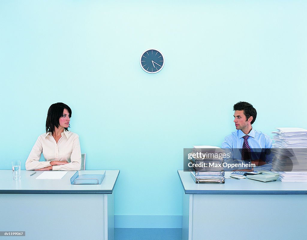 Businesswoman Sitting at a Tidy Desk Looking Sideways at a Businessman Sitting at a Messy Desk