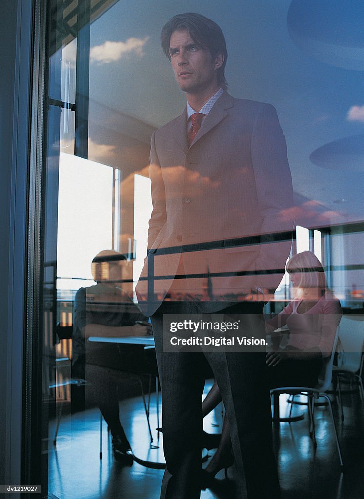 Pensive Businessman Looking Through an Office Window With His Hands in His Pockets and Colleagues Sitting in the Background