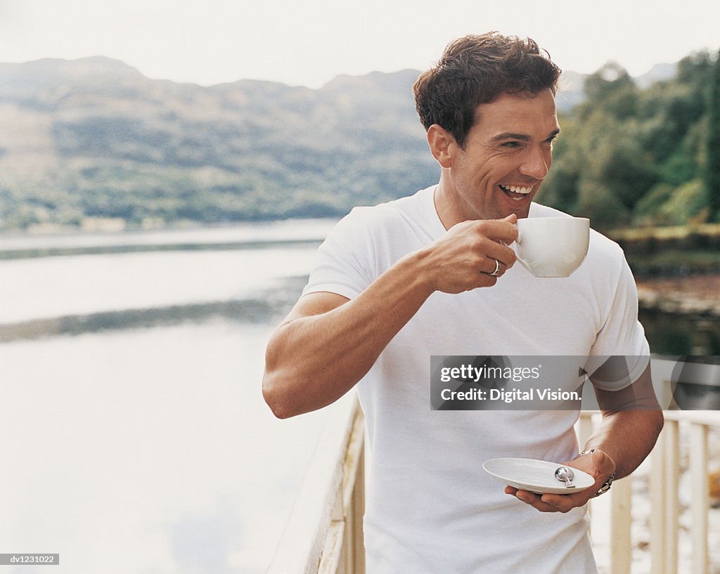 Man Standing Laughing as he Drinks From a Coffee Cup on a Balcony Beside a Lake
