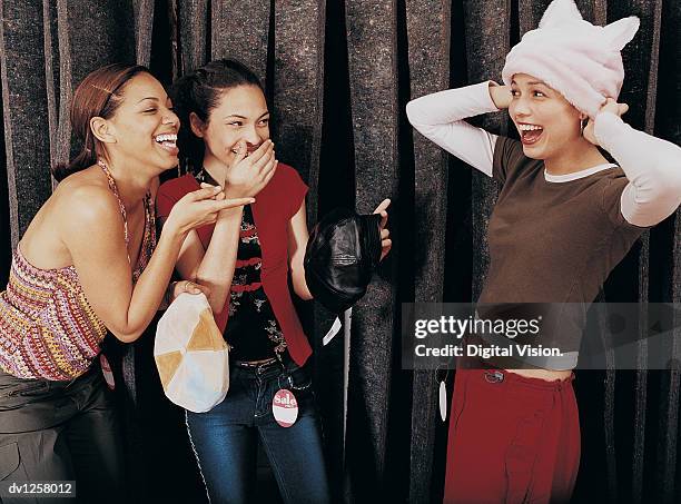 two girls laughing at a young girl trying on a hat in the changing room of a clothes shop - girl changing room shop stock-fotos und bilder