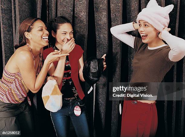 two girls laughing at a young girl trying on a hat in the changing room of a clothes shop - girl changing room shop stock pictures, royalty-free photos & images