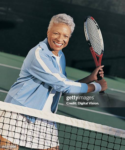 portrait of a mature woman standing behind a net on a tennis court holding a racket - tennis court stock pictures, royalty-free photos & images