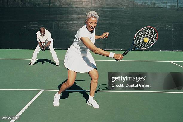 senior woman hitting a tennis ball on a tennis court and a senior man standing behind - tennis woman stockfoto's en -beelden